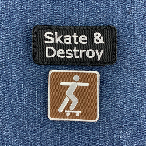 Two embroidered patches displayed on a blue denim fabric background. The top patch is black with white text reading 'Skate & Destroy'. The bottom patch is a brown and white skatepark sign.