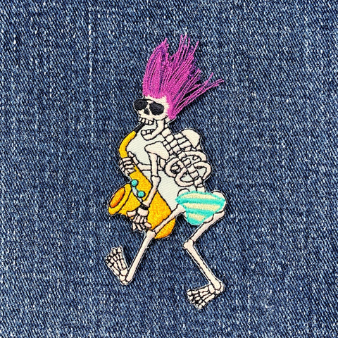 Embroidered patch depicting a walking skeleton playing a yellow saxophone wearing sunglasses, a spiked purple hairdo made of loose thread, and blue and yellow striped shorts, displayed on a denim background