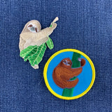 Two embroidered sloth-themed patches on a blue denim background: one irregular patch showing a beige sloth with green leaves, and one circular patch with a brown sloth climbing a green branch inside a blue circle with a yellow border.