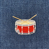 A patch depicting a snare drum with drumsticks on top, primarily red with white and gray accents on a denim background.