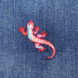A colorful embroidered patch in the shape of a lizard with a geometric pattern on a denim background