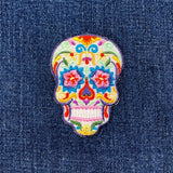 A colorful embroidered patch in the shape of a sugar skull, designed as a Dia de los Muertos or Halloween badge on a denim background