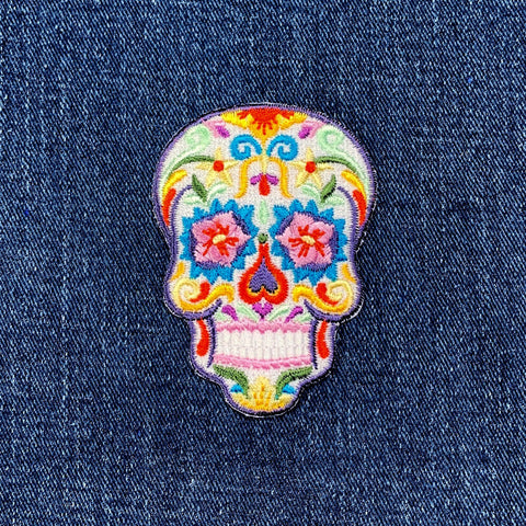 A colorful embroidered patch in the shape of a sugar skull, designed as a Dia de los Muertos or Halloween badge on a denim background
