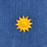 A small embroidered yellow sun patch with a textured center, placed on a blue denim fabric background.