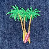 A close-up of an embroidered patch featuring three green palm trees with tan trunks and a pink and yellow surfboard, on a dark blue denim fabric background.