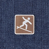 A square embroidered patch featuring a white surfer icon on a brown background, bordered in white and placed on a blue denim fabric.