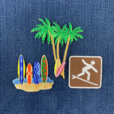 Embroidered beach-themed patches on a blue denim background, featuring green palm trees, colorful surfboards, and a brown square patch with a white surfer silhouette.