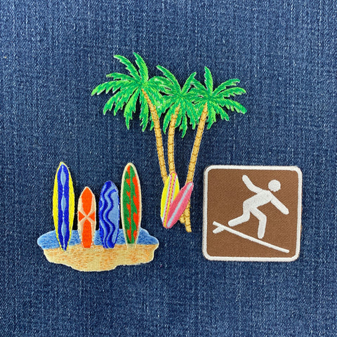 Embroidered beach-themed patches on a blue denim background, featuring green palm trees, colorful surfboards, and a brown square patch with a white surfer silhouette.
