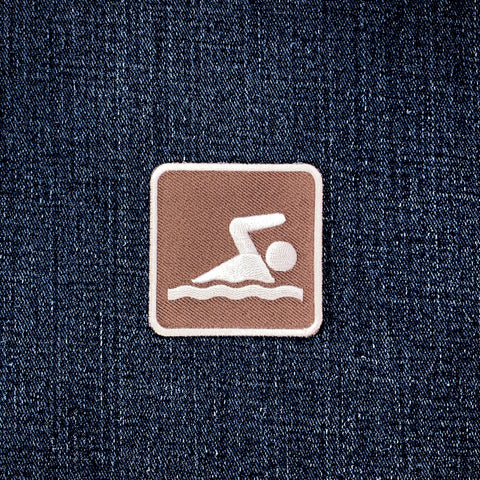 A square brown patch with a white swim icon, displayed on a dark blue denim fabric background.