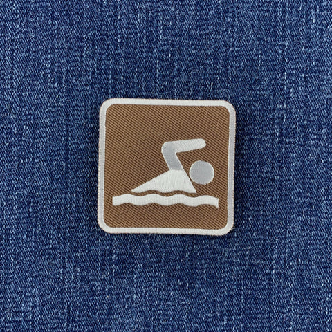 A square embroidered patch featuring a white swimmer icon on a brown background, bordered in white and placed on a blue denim fabric.