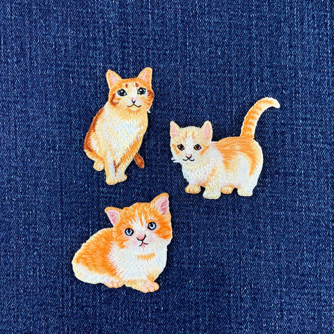 Three embroidered orange and white cat patches on a blue fabric background