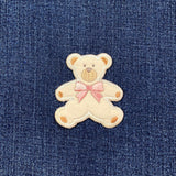 A teddy bear-shaped applique patch with a pink bow detail on the front and displayed on a denim fabric