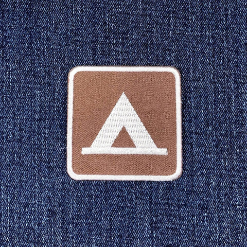 A square brown embroidered patch with a white border, featuring a white stylized tent logo on a blue denim fabric background.
