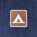 A square brown embroidered patch with a white border, featuring a white stylized tent logo on a blue denim fabric background.