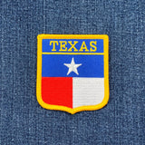 A shield-shaped embroidered patch with the Texas state flag colors and the word 'TEXAS' across the top. The patch has a yellow outer edge, with blue, red, and white fields and a white star.