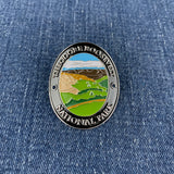 An oval enamel Walking Stick Medallion featuring a landscape of Theodore Roosevelt National Park, displayed on a blue denim fabric background.