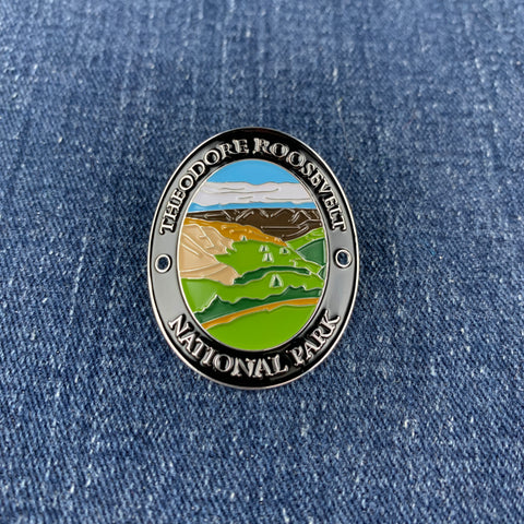 An oval enamel Walking Stick Medallion featuring a landscape of Theodore Roosevelt National Park, displayed on a blue denim fabric background.