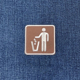 A square brown embroidered patch with a white icon of a person disposing of trash, placed on a blue denim fabric background.