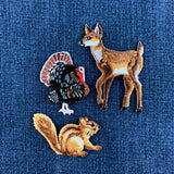 Three embroidered animal patches, including a turkey, fawn, and chipmunk, displayed on a blue denim fabric background.