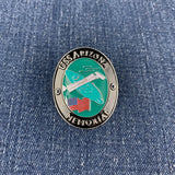 An oval metal commemorative medallion reading 'USS ARIZONA MEMORIAL' with a turquoise center, silver border, and placed on blue denim fabric.