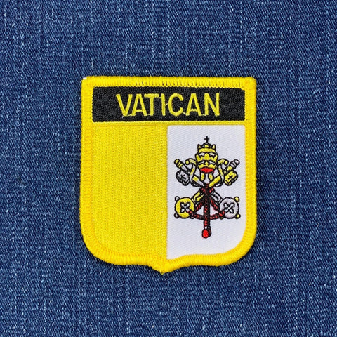 A shield-shaped embroidered patch with a yellow background and the word 'VATICAN' across the top on a denim background.