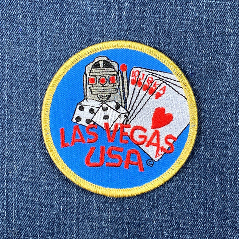 Embroidered circular blue patch with a golden border. Features a slot machine, dice, and a royal flush poker hand over the red text "LAS VEGAS USA" on a denim background