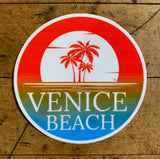 A circular sticker featuring a vibrant orange and blue sunset over water, with a silhouette of three palm trees in front of a large white sun. The text 'VENICE BEACH' is printed in large white letters across the bottom of the design.