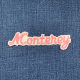 A pink, iron-on embroidered patch that reads 'Monterey' in a cursive font on a denim background