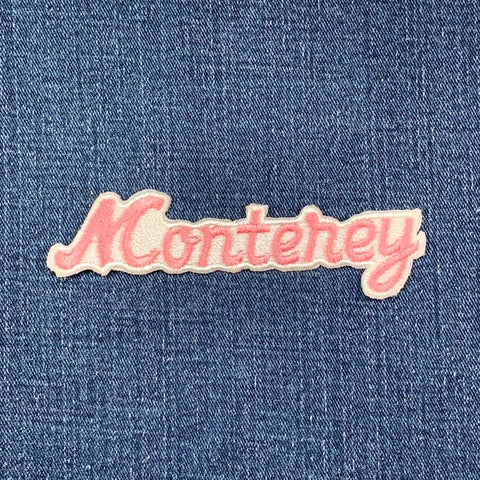 A pink, iron-on embroidered patch that reads 'Monterey' in a cursive font on a denim background