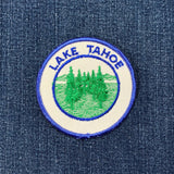 Circular white embroidered patch for Lake Tahoe, depicting a cluster of green evergreen trees against a background of blue water and soft hills on a denim background.