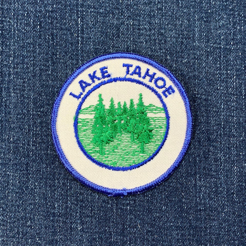 Circular white embroidered patch for Lake Tahoe, depicting a cluster of green evergreen trees against a background of blue water and soft hills on a denim background.