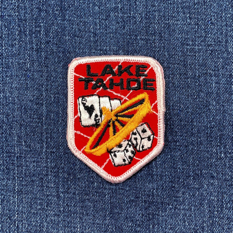 A vintage patch with a red background featuring the text 'LAKE TAHOE' and an illustration of a roulette wheel, playing cards, and dice, displayed on a denim background.