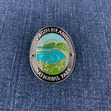 An oval enamel Walking Stick Medallion featuring a landscape of Virgin Islands National Park with turquoise water, green hills, and palm trees, bordered by a black ring with white text, shown on a blue denim background.