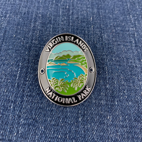 An oval enamel Walking Stick Medallion featuring a landscape of Virgin Islands National Park with turquoise water, green hills, and palm trees, bordered by a black ring with white text, shown on a blue denim background.
