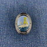 An oval enamel Walking Stick Medallion featuring a Voyageurs National Park design with a yellow sailing ship on a rocky island, set against blue water and sky, resting on a blue denim background.