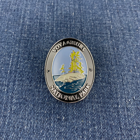 An oval enamel Walking Stick Medallion featuring a Voyageurs National Park design with a yellow sailing ship on a rocky island, set against blue water and sky, resting on a blue denim background.