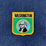 A shield-shaped embroidered patch with the text 'WASHINGTON' at the top, a portrait of George Washington in the center, displayed on a denim background.