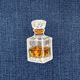 A small embroidered patch of a decanter with a clear glass body, amber liquid inside, and a tan stopper, centered on a blue denim-like textured background.