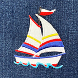 A colorful embroidered patch depicting a sailboat with red, blue, and yellow sail shades, on a denim background.