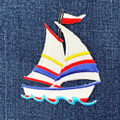 A colorful embroidered patch depicting a sailboat with red, blue, and yellow sail shades, on a denim background.