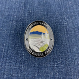 An oval enamel Walking Stick Medallion featuring a landscape design of White Sands National Park, with a silver-toned border and colorful center, placed on a blue denim background.