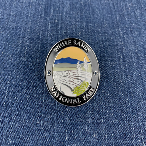 An oval enamel Walking Stick Medallion featuring a landscape design of White Sands National Park, with a silver-toned border and colorful center, placed on a blue denim background.