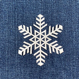White embroidered snowflake patch with intricate six-pointed design against a deep blue, textured denim fabric background.