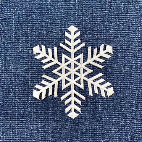 White embroidered snowflake patch with intricate six-pointed design against a deep blue, textured denim fabric background.