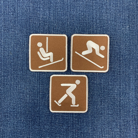 Three square brown patches with white winter sports icons, including ice skating, downhill skiing, and a chairlift, arranged on a blue denim fabric background.