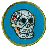 A patch featuring a stylized sugar skull design on a blue background with a green border.