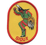 An oval-shaped embroidered patch featuring a Native American figure with the text 'SIoux' below. The patch has a red border and displays a figure in green attire with decorative elements and a red backdrop.