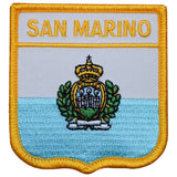 A shield-shaped embroidered patch with the words 'SAN MARINO' at the top and an emblem with a crown, laurel wreath, and banner in the center. The patch has a yellow border and a light blue background.